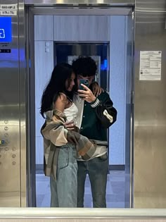 a man and woman standing in front of an elevator looking at their cell phones together