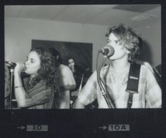 black and white photograph of two people singing into microphones while another person plays guitar in the background