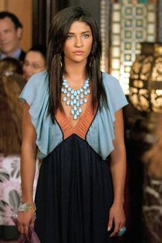 a woman with long hair wearing a black dress and blue necklace, standing in front of a group of people