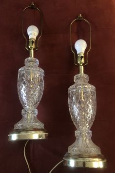 two clear glass lamps are sitting on a brown tableclothed surface, one is turned off and the other has a cord attached to it