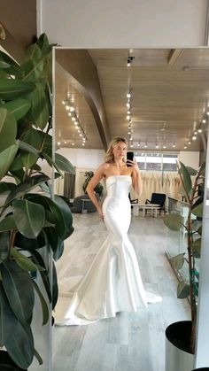 a woman in a white dress taking a selfie with her cell phone while standing next to a plant