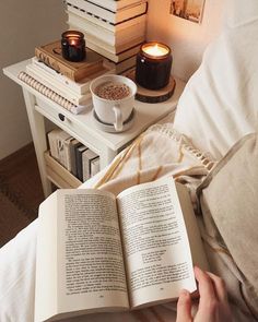 a person reading a book in bed with candles on the side table and books stacked up behind them