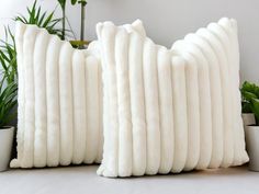 two white pillows sitting next to each other on top of a table with plants in the background