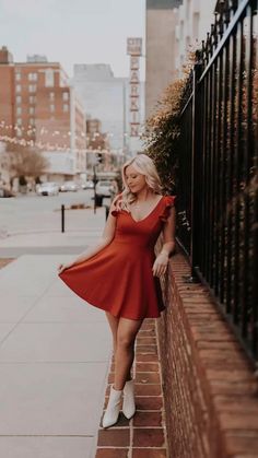 a woman leaning against a fence wearing a red dress and white boots, posing for the camera