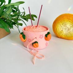 a pink cup with two needles in it next to a potted plant and ball of yarn