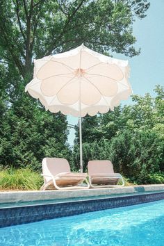 two lounge chairs under an umbrella next to a swimming pool with trees in the background