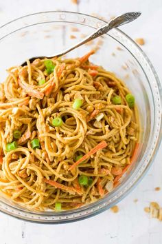 a glass bowl filled with noodles, carrots and green onions next to a fork