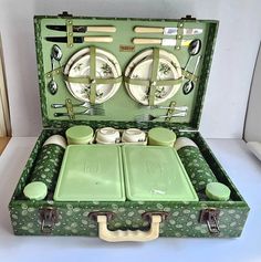 an old green suitcase with plates and utensils in the inside, sitting on a white surface