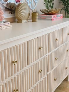 a white dresser topped with lots of drawers and gold knobs next to a plant