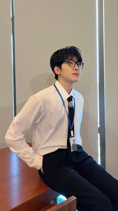 a young man sitting on top of a wooden desk wearing glasses and a tie with his hands on his hips