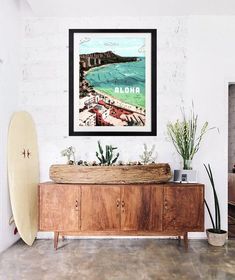 a surfboard sitting on top of a wooden cabinet next to a painting and potted plants