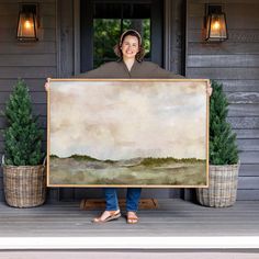 a woman holding up a large painting in front of a house with potted plants