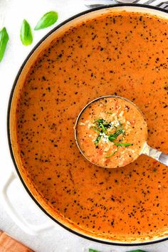 a bowl of tomato soup with a spoon in it and basil leaves on the side