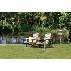 two wooden chairs sitting on top of a grass covered field next to a lake and trees