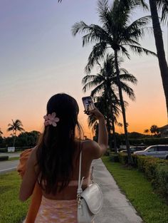 a woman is taking a photo with her cell phone at sunset on the sidewalk in front of palm trees