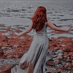 a woman with red hair is standing on rocks near the water and holding her arms out