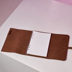 an open brown leather notebook sitting on top of a white table next to a pink wall