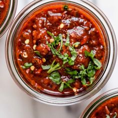 three jars filled with chili sauce and garnished with fresh herbs on top, sitting on a marble surface