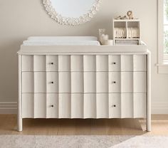 a white dresser with drawers and a baby crib in front of a round mirror