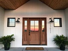 two potted plants sit on the front porch of a white house with wood paneling