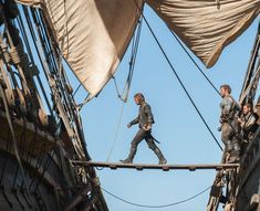 two men walking across a rope bridge on top of a tall ship in the ocean