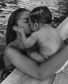 a woman holding a baby in her arms while sitting on a dock next to the water