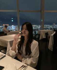 a woman sitting at a table with a wine glass in her mouth and looking out the window