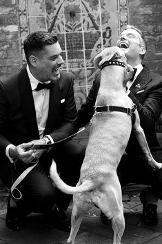 two men in tuxedos are sitting on a bench and petting a dog