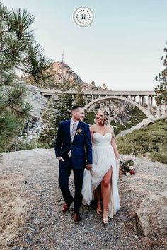 a bride and groom standing on top of a mountain with a bridge in the background