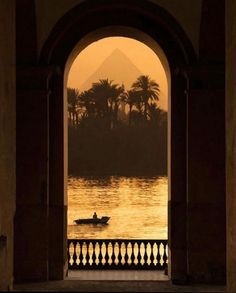 a person in a small boat on the water near an arch with a view of mountains and palm trees