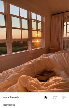 an unmade bed with a book on it in front of two large windows at sunset