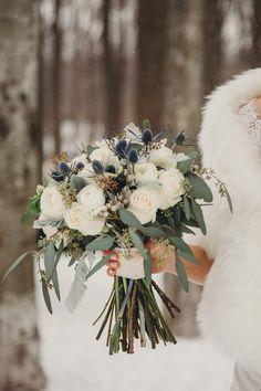 a bride holding a bouquet of white flowers and greenery in her hand while standing in the snow