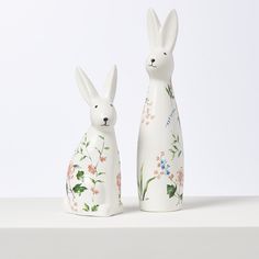 two white rabbits sitting next to each other on top of a shelf with flowers and leaves painted on them