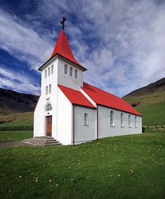 a white church with a red roof and steeple on the grass in front of mountains