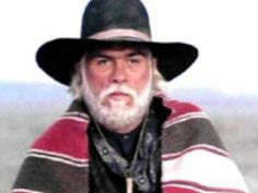 an old man wearing a black hat and striped blanket is looking at the camera while standing in front of him