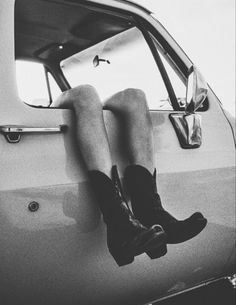 black and white photograph of a person sitting on the hood of a car with their legs hanging out