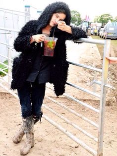 a man in a furry coat drinking from a drink cup while standing next to a fence