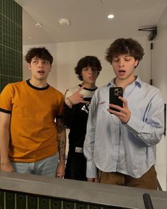 three young men standing in front of a mirror looking at their cell phones and making faces