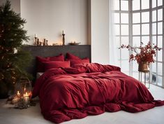 a bed with red sheets and pillows next to a christmas tree in front of a window