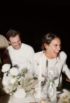 a man and woman sitting at a table with flowers in front of them, laughing