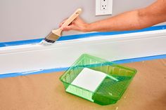 someone using a paint brush to paint the trim on a wall with blue tape and a green container