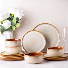 a white vase filled with flowers next to plates and cups on a counter top in front of a wall