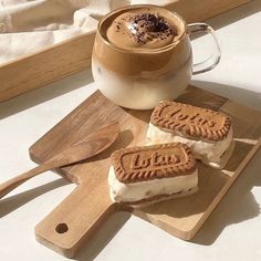 two cookies are on a cutting board next to a cup of hot chocolate and a spoon