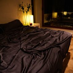 an unmade bed in front of a window with candles on the floor next to it