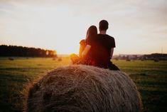 two people sitting on the back of a hay bale in a field at sunset