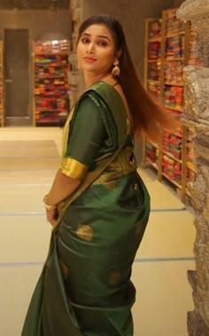 a woman in a green and gold sari standing next to a stone wall with stacks of books behind her