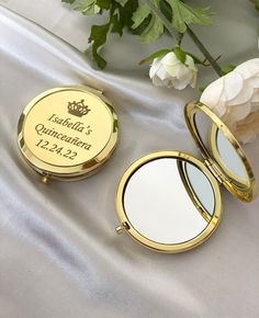 two compact mirrors sitting next to each other on a white satin covered surface with flowers in the background