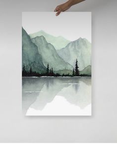 a person holding up a card with watercolor painting on it and mountains in the background