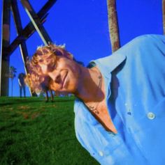 a man with red hair and blue shirt standing in front of palm trees on a sunny day