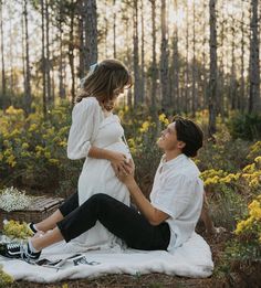 a pregnant woman sitting on top of a blanket next to a man in the woods